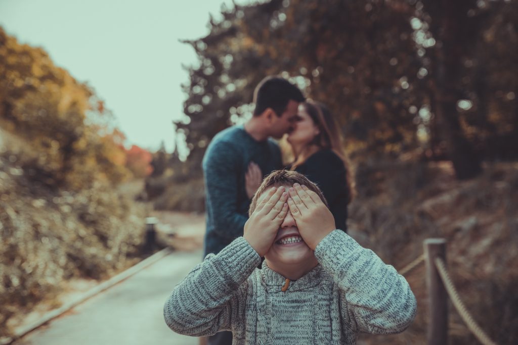 Kind hält sich die Augen zu während die Eltern im Hintergrund knutschen